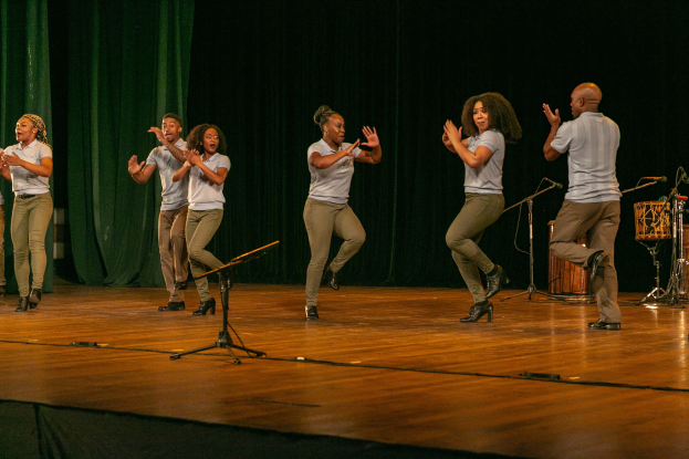 Eine Gruppe von Menschen auf einer Bühne, die klatschen und feiern, mit Mikrofonen, Instrumenten und einem Vorhang im Hintergrund, wahrscheinlich ein Tanzteam, das seinen Erfolg anerkennt.