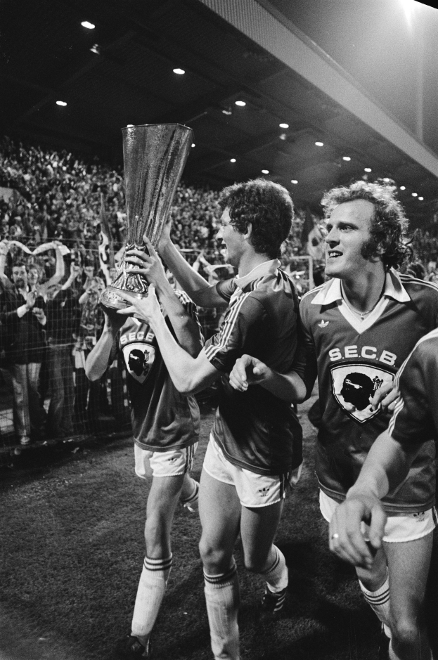 Schwarzes Bild von Männern, die einen Fußballsieg feiern, indem sie eine Trophäe hochhalten, auf einem Feld, mit einer Menge hinter einem Zaun und überdachter Stadionbeleuchtung.