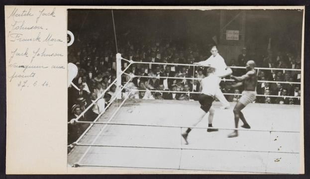Schwarz-weiß-Foto von zwei Boxern, Jack Johnson und Frank Morgan, in einem Boxring mit Zuschauern und einer Wand im Hintergrund.