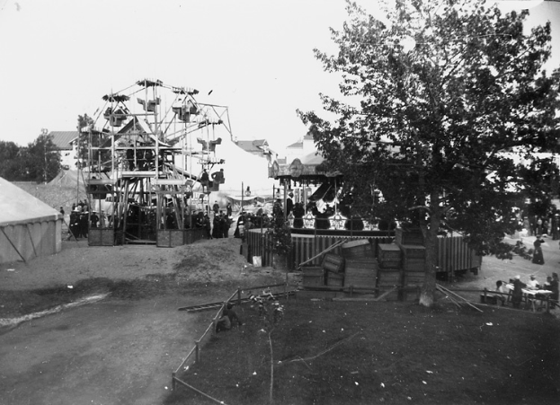 Ein Schwarz-Weiß-Foto eines Karnevals mit einem Riesenrad in der Mitte, umgeben von Menschen, Zelten, Bäumen und anderen Gegenständen, mit einer Straße am unteren Bildrand.