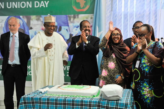Eine Gruppe von Menschen, die um einen Tisch mit einer Torte, Tellern und anderen Gegenständen stehen und klatschen, mit einem Banner, Vorhängen und einer Wand im Hintergrund.