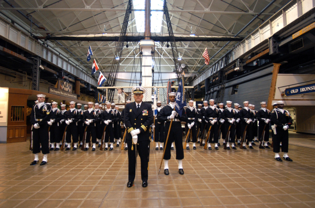 Eine Gruppe uniformierter Männer in einem großen Raum mit Gewehren, Flaggen, Schildern und Deckenleuchten und Stangen.
