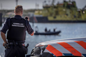 Ein Polizist in Uniform steht neben einem Polizeiwagen in der Nähe eines Gewässers und hält ein Funkgerät in der Hand, mit einem Boot im Hintergrund, das ein paar Menschen trägt, und dem Himmel darüber.
