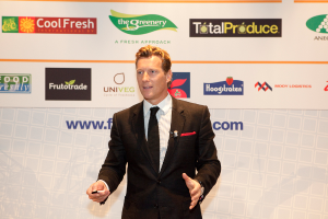 Ein Mann in einem schwarzen Blazer steht und hält ein Objekt, während er spricht, mit einem weißen Banner und einem weißen Brett mit organisationalen Logos im Hintergrund.