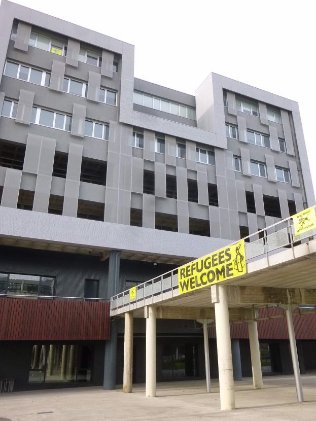 Großes Gebäude mit einem "Willkommen bei den Flüchtlingen"-Schild, umgeben von Säulen und einem Geländer, vor einem Himmel-Hintergrund.