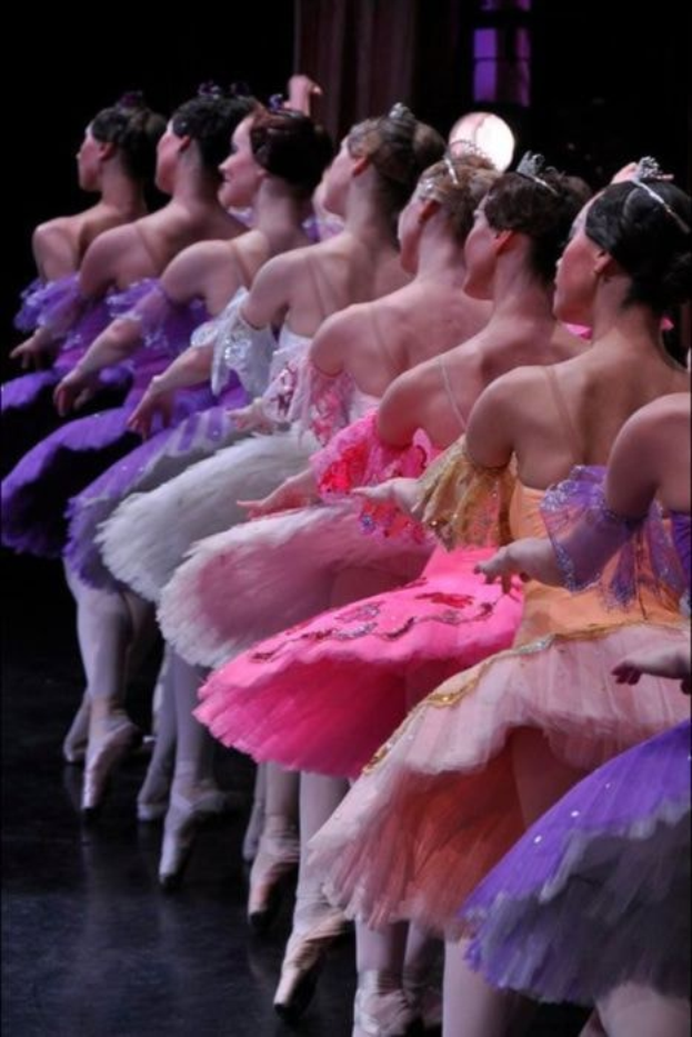 Eine Gruppe von Balletttänzerinnen in rosafarbenen Tutus sitzt auf einer Bühne, beleuchtet von einem Licht im Hintergrund, ihre synchronen Bewegungen schaffen eine fesselnde Szene.