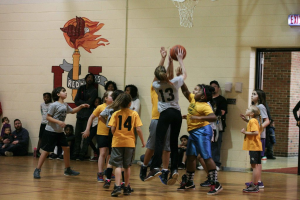 Kinder in gelben und grauen T-Shirts spielen Basketball, wobei ein Mädchen in einem grauen T-Shirt mit der Nummer 13 einen Wurf versucht. Im Hintergrund sitzt ein Mann in einem blauen Hemd in der Nähe einer weißen Wand mit Text, und rechts davon ist ein Ausgangsschild und ein rotes Backsteingebäude zu sehen.