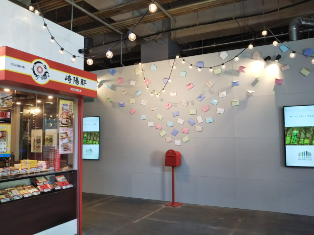 Ein Ladeneinrichtungsbild mit einer Vitrine voller verschiedener Lebensmittel, einem roten Briefkasten auf dem Boden, einem Anzeigebildschirm an der Wand mit einem Yokohama Kitchen-Schild, Papieren an den Wänden, Deckenleuchten und sichtbaren Rohren.