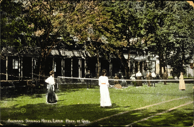 Schwarz-weißes Foto von Menschen, die Tennis auf dem Rasen vor dem Abenakis Springs Hotel Lawn in Provo, Quebec, spielen, mit einem Netz, Stühlen, Bäumen und einem Gebäude im Hintergrund.