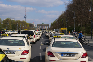 Eine lange Reihe von Taxis, die an einer belebten Straße in Berlin, Deutschland, geparkt sind, mit Fußgängern und Radfahrern in der Nähe, flankiert von Laternenmasten und Bäumen und Gebäuden, einem Bogen und einem Turm im Hintergrund unter einem bewölkten Himmel.