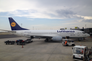 Ein Lufthansa Airbus A320-200 steht am Frankfurter Flughafen, umgeben von diversen Bodenfahrzeugen, Wagen, Verkehrskegeln, Pfosten und Texttafeln unter einem bewölkten Himmel.