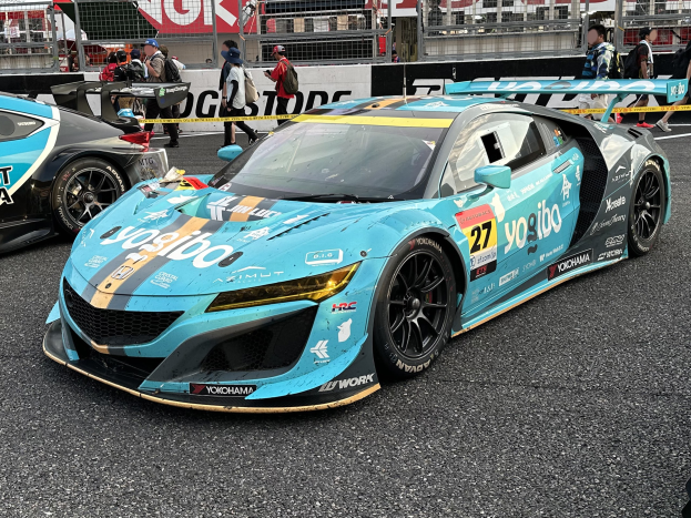Gruppe von Autos auf einer Rennstrecke geparkt, mit Menschen in Mützen und Taschen im Hintergrund, Rennstrecke Umgebung einschließlich Texttafeln, Geländer, Pfosten und ein fernes Gebäude, mit "Yokohama Acura NSX GT3" Text unten.