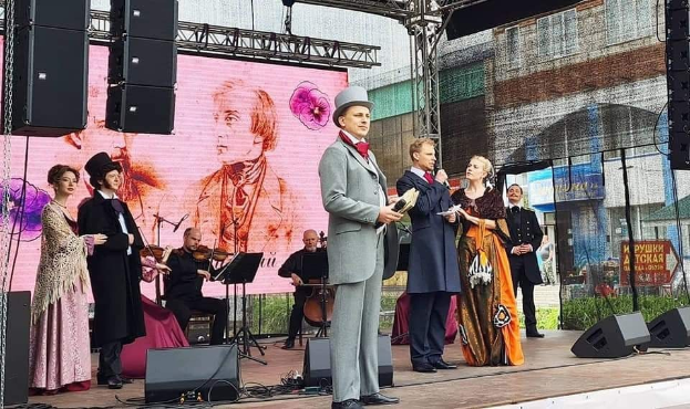 Eine Gruppe von Menschen auf einer Bühne bei einem Event, mit einem Mann in der Mitte mit Mikrofon und Buch, einer Frau links von ihm auf einem Stuhl mit einem Musikinstrument und einem Mann rechts von ihm mit einem Papier in der Hand, während im Hintergrund ein Geigenspieler sitzt und ein Ständer mit einem Buch vor ihm zu sehen ist.