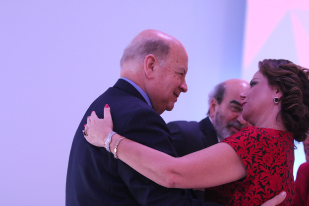 Zwei Menschen umarmen sich herzlich auf einer Bühne, umgeben von anderen; der Mann trägt ein Armband.