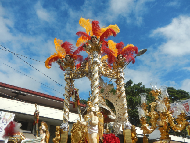 Ein mit goldenen und roten Federn geschmückter Karnevalswagen, der Menschen trägt, wird in einem Umzug mit einem Gebäude, Bäumen, Drähten und einer Fahne in der Nähe unter einem sichtbaren Himmel gezeigt.