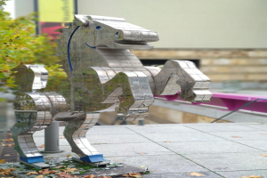 Eine Metall-Pferdeskulptur steht auf einem Gehweg umgeben von trockenen Blättern, mit einem Baum links und einer Wand und einem Objekt im Hintergrund.