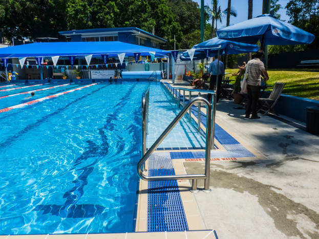 Großer Freibad mit mehreren Schwimmern, umgeben von Geländern, Sonnenschirmen, Liegestühlen, Tischen, Grünflächen und einem Gebäude unter einem klaren blauen Himmel.