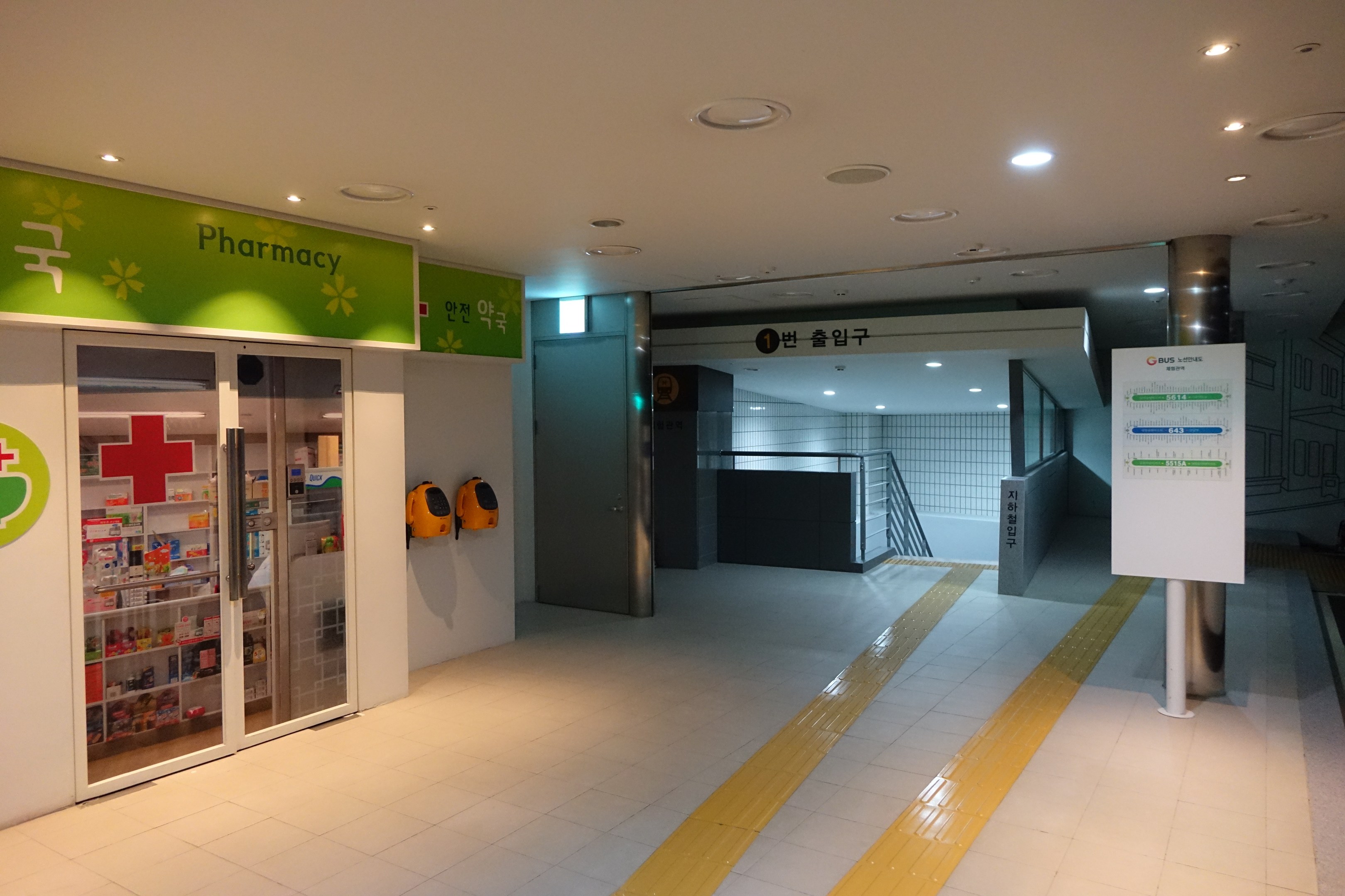 Eingang zu einer Apotheke mit einem "Apotheke"-Schild auf der linken Seite, einer Tafel mit Text auf der rechten Seite, beleuchteten Lampen oben und undeutlichen Hintergrundobjekten.