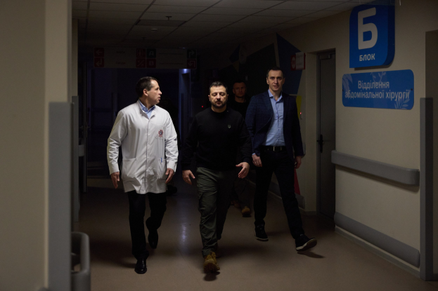 Eine Gruppe von Männern, die einen hell erleuchteten Krankenhausflur entlanggehen, mit einer Tür auf der rechten Seite und Texttafeln auf der linken Seite, die besorgt aussehen.