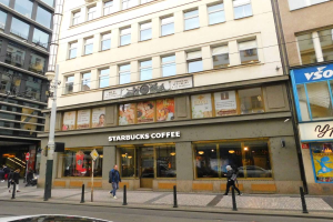 Ein Starbucks-Kaffee-Shop an einer Straßenecke in Berlin, Deutschland, mit Menschen auf dem Gehweg, einem Auto auf der Straße und einem Mast mit Drähten entlang der Straße. Das Gebäude hat Fenster und Schilder mit Text und Bildern.