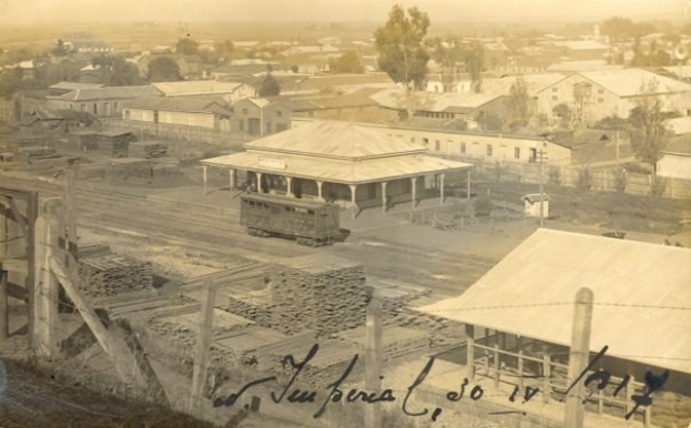 Ein altes Schwarz-Weiß-Foto einer Stadt mit einem Zug auf den Schienen, umgeben von Gebäuden, Bäumen, Pfählen und einem Zaun, mit Text unten.