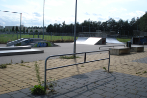 Ein Skatepark mit Rampen, Geländern, Grünflächen, Pfählen, Bäumen, Gebäuden und einem bewölkten Himmel im Hintergrund.