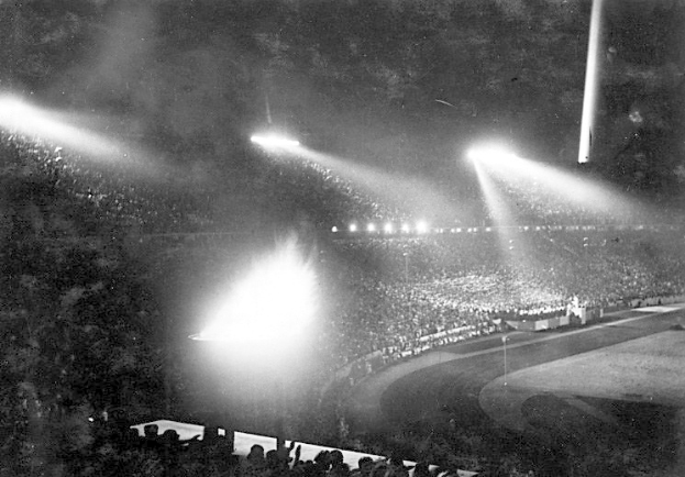 Schwarzes und weißes Foto eines überfüllten Stadions, das von Flutlicht beleuchtet wird, vor dunklem Hintergrund.