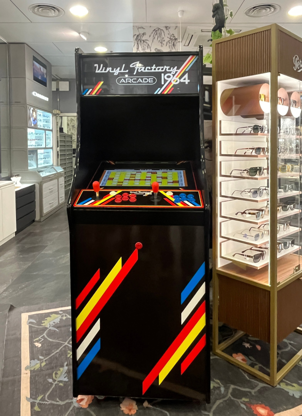 Ein Ladeneinrichtungsbild mit einem zentralen Vintage-Arcade-Automaten, einem Glasvitrinenschrank auf der rechten Seite und Retro-Stil-Beleuchtung mit Schäben, einem Fernseher und anderen Gegenständen im Hintergrund.