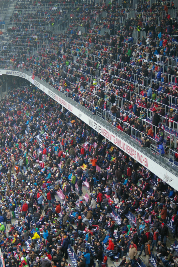 Eine große Menschenmenge sitzt und steht auf den Tribünen eines Stadions mit Geländern und Texttafeln im Hintergrund.