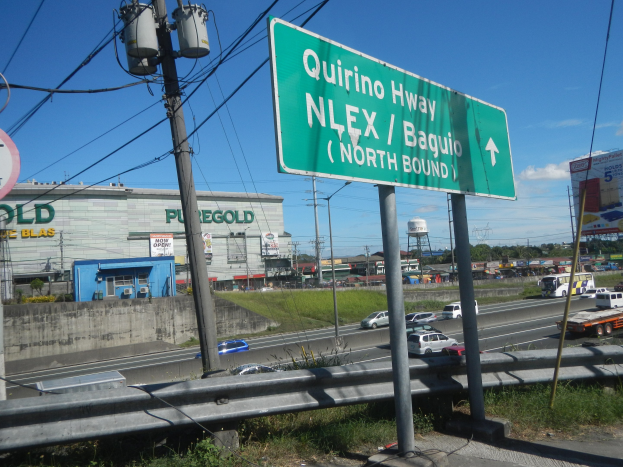 Grüne Highway-Schilder neben einer Straße mit Fahrzeugen, Strommasten, Gras, Gebäuden, Bäumen und einem klaren blauen Himmel.