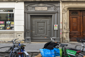 Mehrere Fahrräder stehen in einer Reihe auf dem Gehweg vor einem Gebäude mit einer Tür und einem Fenster auf der linken Seite.