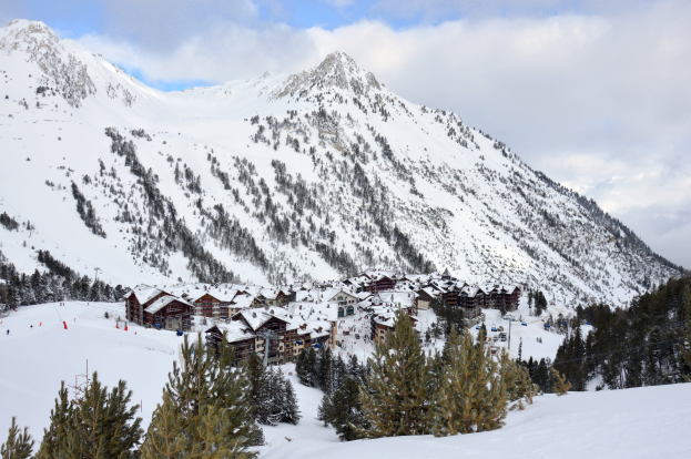 Eine Winterlandschaft in einem Skigebiet mit schneebedeckten Bergen im Hintergrund, schneebeladenen Bäumen unten und einer Hütte zwischen den Bäumen sowie einer bewölkten Himmel.