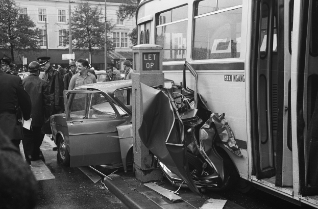 Schwarz-weißes Bild eines Busses, der auf der Seite der Straße in ein Auto gekracht ist, mit Menschen, die um das Wrack herumstehen und Bäumen und Gebäuden im Hintergrund.