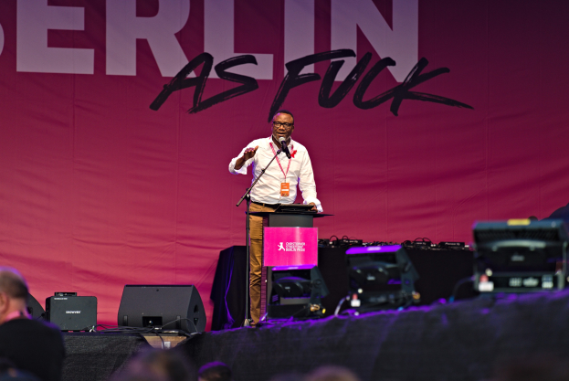Mann in weißem Hemd und braunen Hosen, der in ein Mikrofon spricht, vor einem pinken 'Berlin as Fuck'-Plakat auf einer Bühne mit Blick auf eine Menschenmenge.