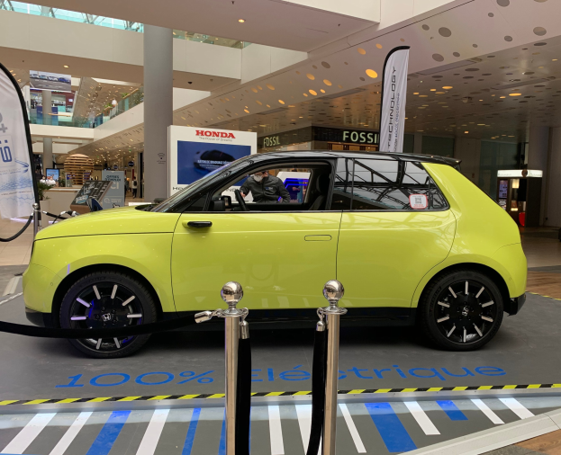 Ein gelbes wasserstoffbetriebenes Elektroauto vor einem Gebäude geparkt, umgeben von Absperrpfosten mit Seilen, Bannern, einer Person im Auto, einer Säule, einer Gruppe von stehenden Menschen, Schildern, einer Topfpflanze, Deckenleuchten und einer Decke mit Lüftern.