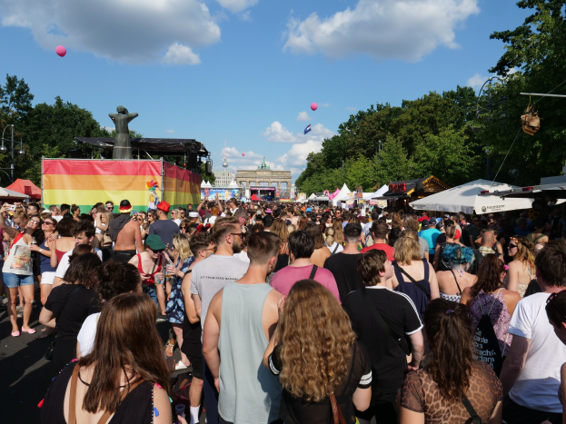Eine gro├če Menge geht eine Stra├če mit Zelten, B├Ąumen, Masten, Lichtern und einer Statue entlang, mit Geb├Ąuden und einem bew├Âlktem Himmel mit Ballons im Hintergrund beim Berlin Pride Festival.