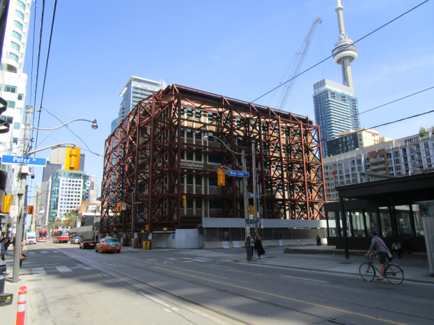 Eine belebte Stadtstraße mit einem großen Gebäude im Hintergrund, Fahrzeugen, einem Radfahrer, Fußgängern, Ampeln, Pfählen mit Schildern, einem Kran und dem Himmel.