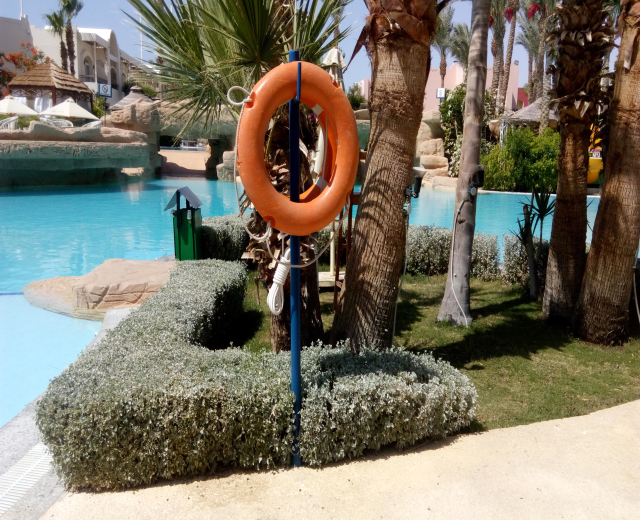 Ein Schwimmbad mit einem Rettungsring in der Mitte, umgeben von Grün, mit Gebäuden, Sonnenschirmen und einem klaren blauen Himmel im Hintergrund.