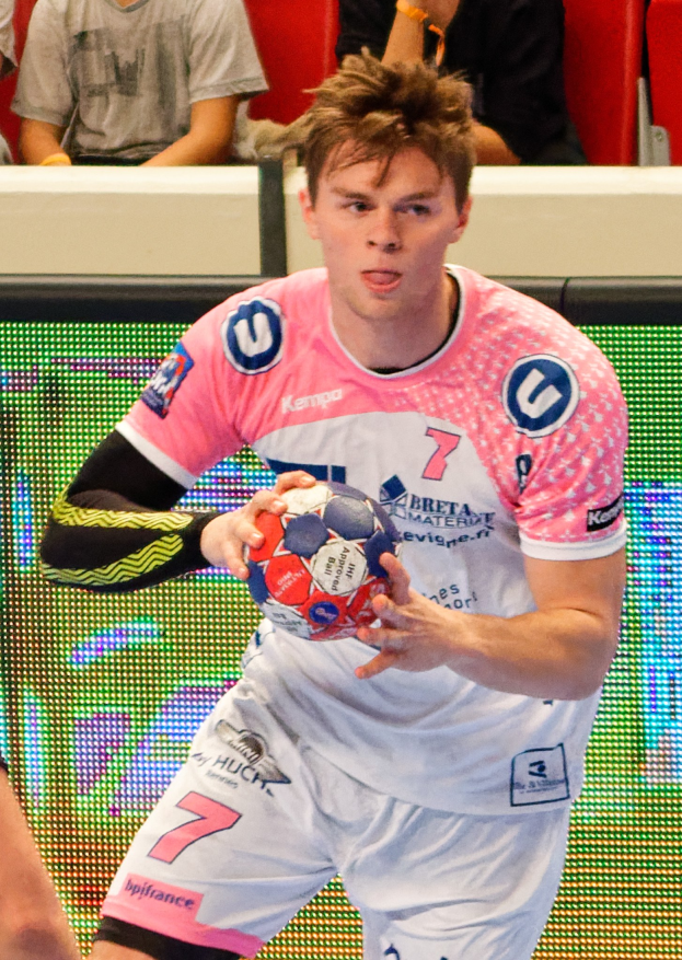 Ein Mann in einem pinken Hemd, der einen Fußball hält, mit der Hand einer anderen Person auf der linken Seite und Menschen im Hintergrund auf Stühlen sitzend.