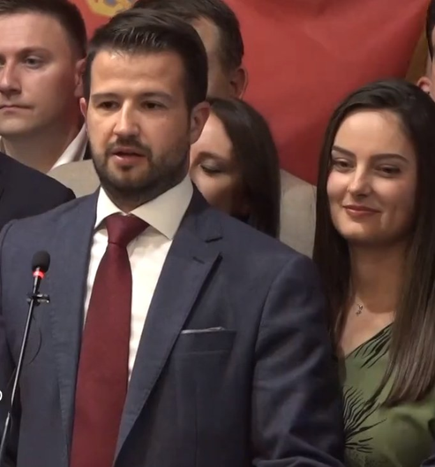 Ein Mann im Anzug spricht in ein Mikrofon auf einem Podium, mit einem Banner dahinter und einer Menge davor, wahrscheinlich zu einem Regierungssozialmedienverbot.