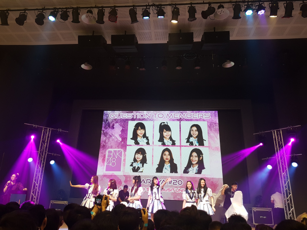 Eine Gruppe von Menschen tritt auf einer hell erleuchteten Bühne mit Trägern auf, während die Menge darunter das Ereignis aufnimmt; ein großer Bildschirm im Hintergrund zeigt Bilder der K-Pop-Gruppe BK48.