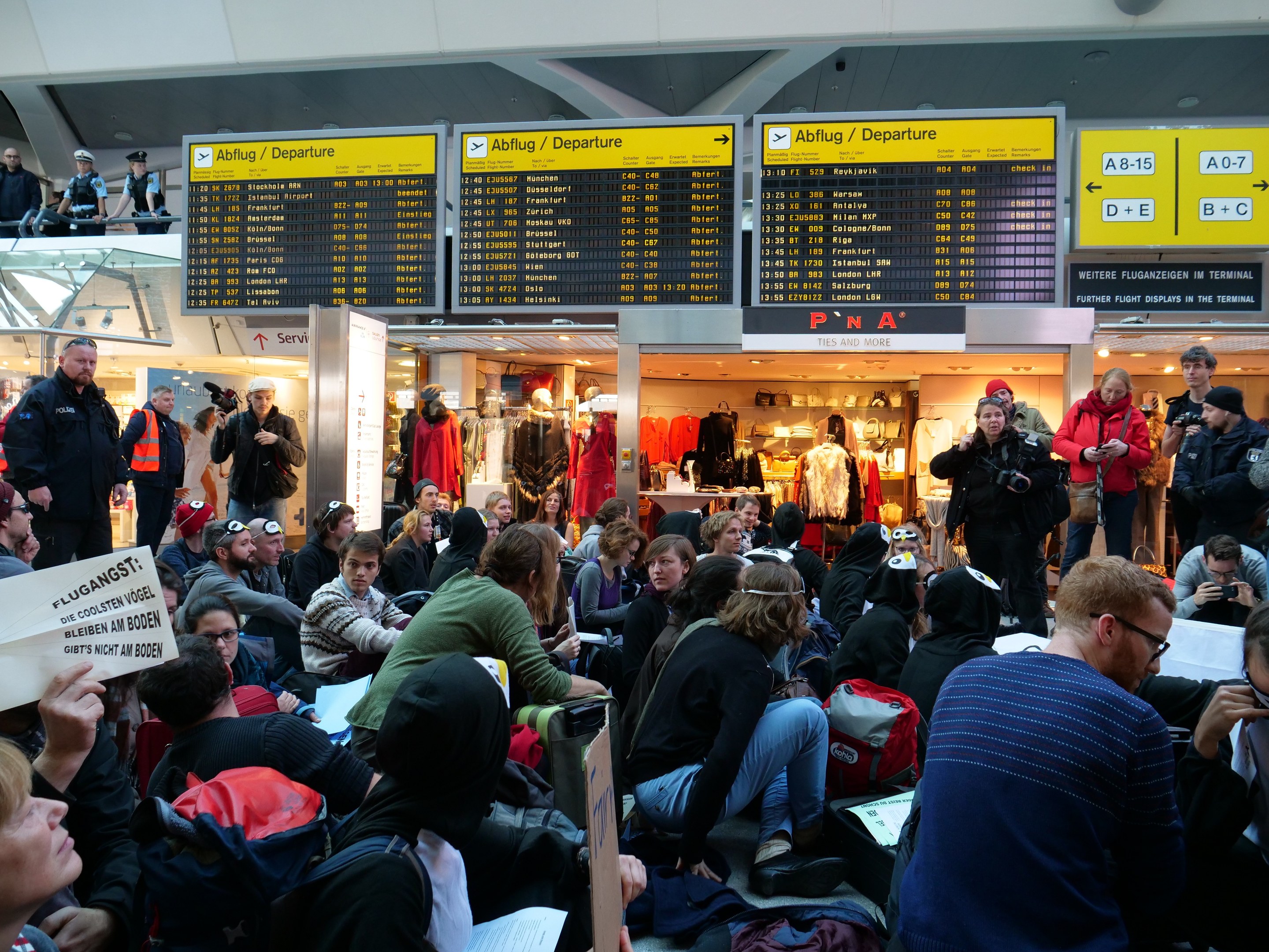 Menschen sitzen und stehen in einem Flughafen während einer Protestaktion, mit Schildern, Puppen und Deckenleuchten im Hintergrund.