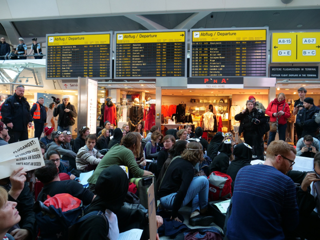 Menschen sitzen und stehen in einem Flughafen während einer Protestaktion, mit Schildern, Puppen und Deckenleuchten im Hintergrund.