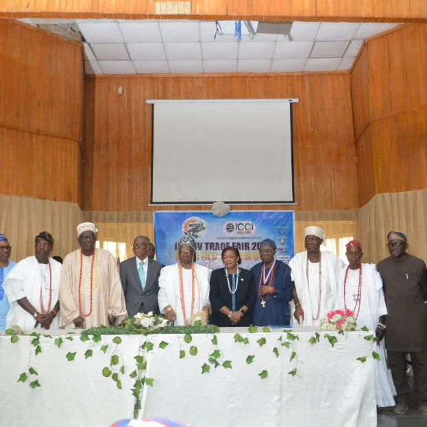 Gruppe von Menschen um einen weißgedeckten Tisch versammelt, der mit Blumen und Blättern geschmückt ist, auf der ICCI International Trade Fair 2019, mit einem Banner, Projektionsbildschirm und Deckenbeleuchtung im Hintergrund.