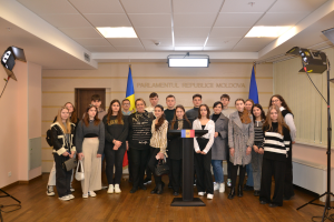 Eine Gruppe von Menschen vor einem Podium mit Mikrofon, umgeben von Fahnen, einer Wand mit dem Text "Moldova Youth Congress 2018", einer Tür, einem Dach mit Deckenleuchten und Ständen mit Leuchten.