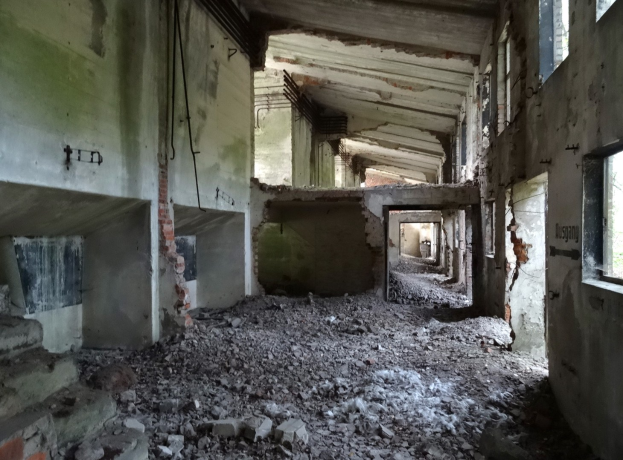 Altes Gebäude in starkem Verfall mit kaputten Fenstern, verstreutem Schutt, freiliegenden Rohren und Schutt auf dem Boden und an den Wänden.