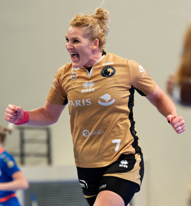 Eine Frau in braunem T-Shirt und schwarzen Shorts rennt auf einem Futsalplatz, feiert mit triumphaler Miene, während zwei andere Frauen im Hintergrund sichtbar sind.