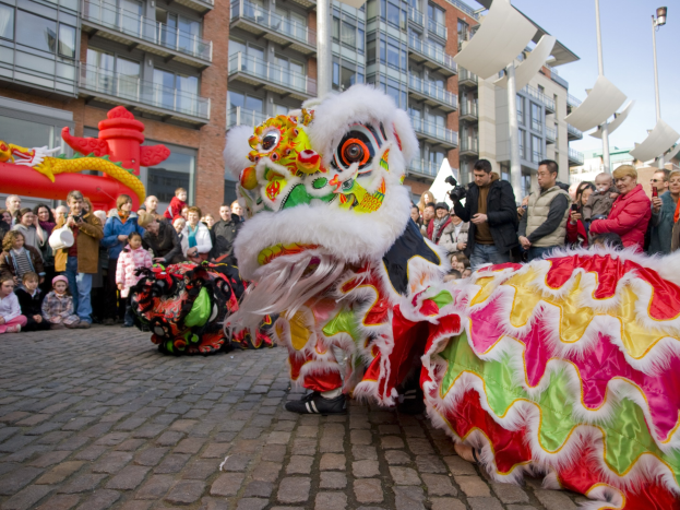 Eine Löwen-Tanzvorstellung während eines chinesischen Neujahrsfestes in Amsterdam, mit einer Zuschauermenge und Menschen, die den Moment mit Kameras festhalten, vor einer Kulisse aus Gebäuden und einem klaren blauen Himmel.