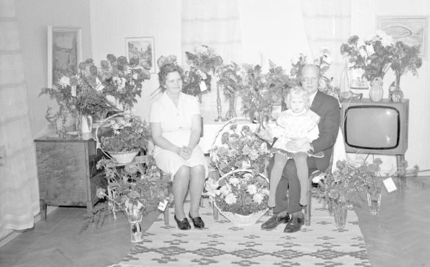 Schwarzes und weißes Foto einer Familie aus drei Personen - einem Mann, einer Frau und einem Kind - auf einem Sofa in einem Wohnzimmer mit Blumenvasen, einem Teppich, gerahmten Wanddekorationen und Vorhängen.
