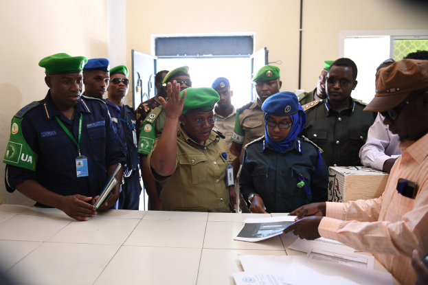 Eine Gruppe von Menschen steht um einen Tisch mit Papieren, einige tragen Mützen und ID-Karten, während eine Person ein Mobiltelefon hält, in einem Raum mit Türen und Fenstern.
